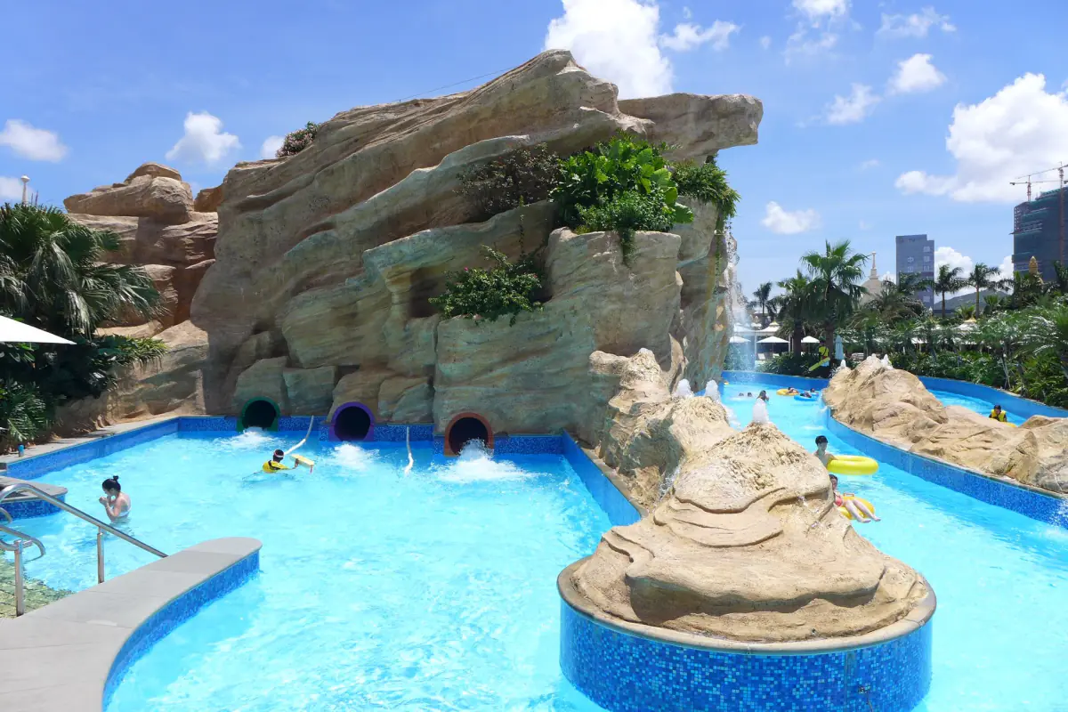 Swimmers drifting on rings through the blue channels of the Grand Resort Deck at Galaxy Macau, past artificial sandstone cliffs with slide mouths, under a sky of broken cloud