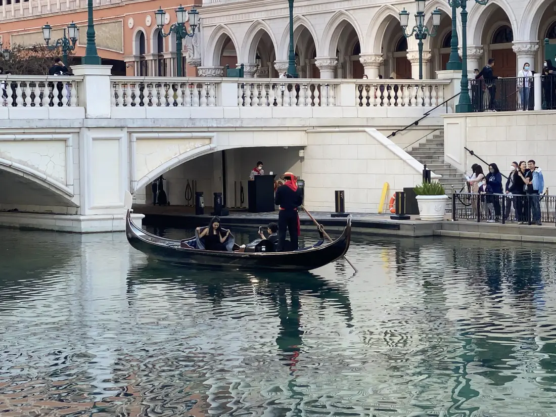 A gondolier in a striped shirt poling a gondola with three passengers along a canal at The Venetian Macao, watched by people leaning on the balustrade above