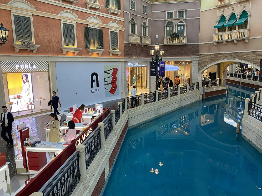 Shoppers on the paved bank of an indoor canal at the Shoppes at Venetian, painted palazzo facades and shopfronts rising on both sides under an artificial sky