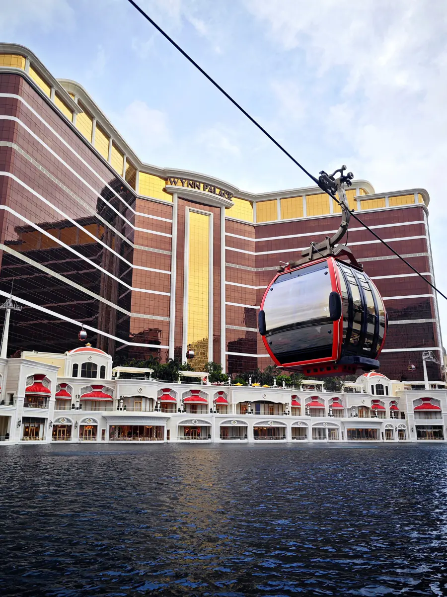 A red SkyCab cable car hanging from its wire above the water of the Performance Lake, with the bronze and gold facade of Wynn Palace filling the sky behind it